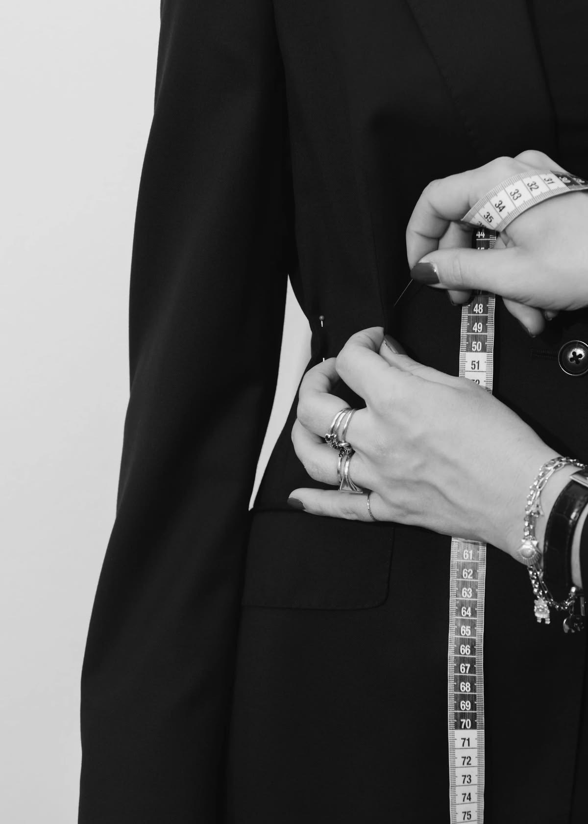 Hands pinning a black jacket at the waist, a tailor’s tape hanging beside them.